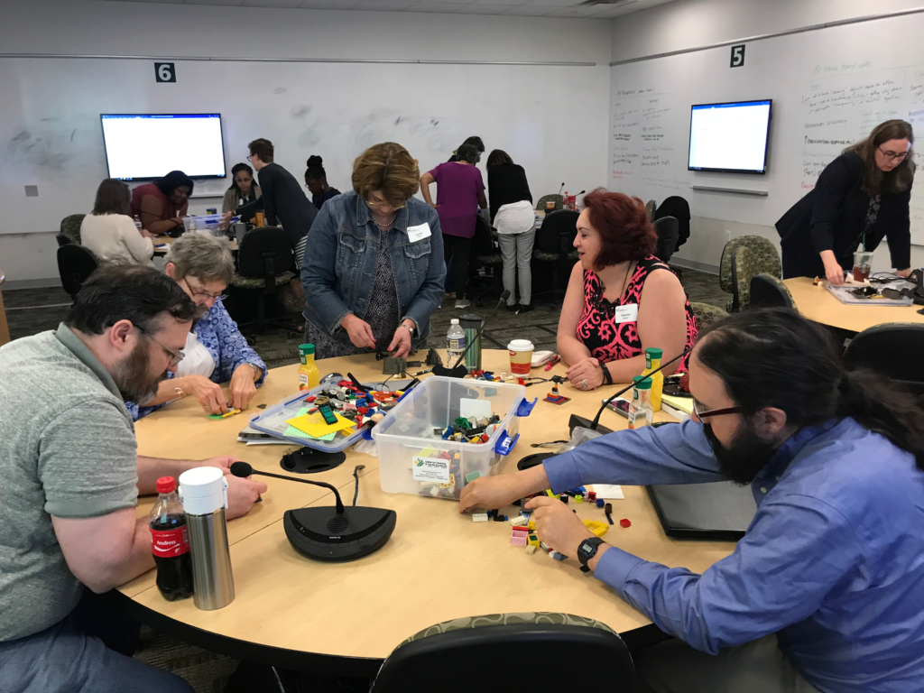 Adults wearing nametags sit around a table playing with legos. The surrounding classroom space has whiteboards with writing on them. The photo implies an interactive conference session.
